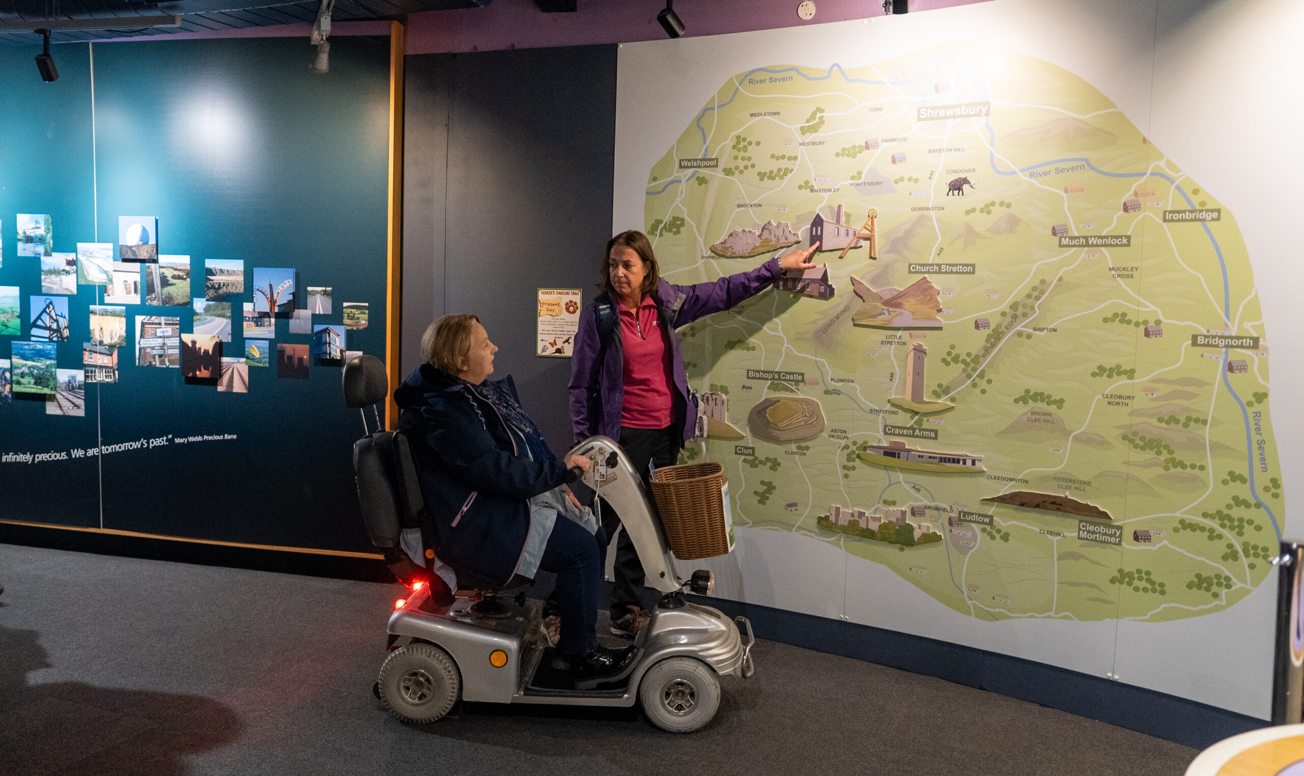 Two women are in an exhibition gallery. One is in a motorised wheelchair and the other is standing Pointing to a large map on the wall.