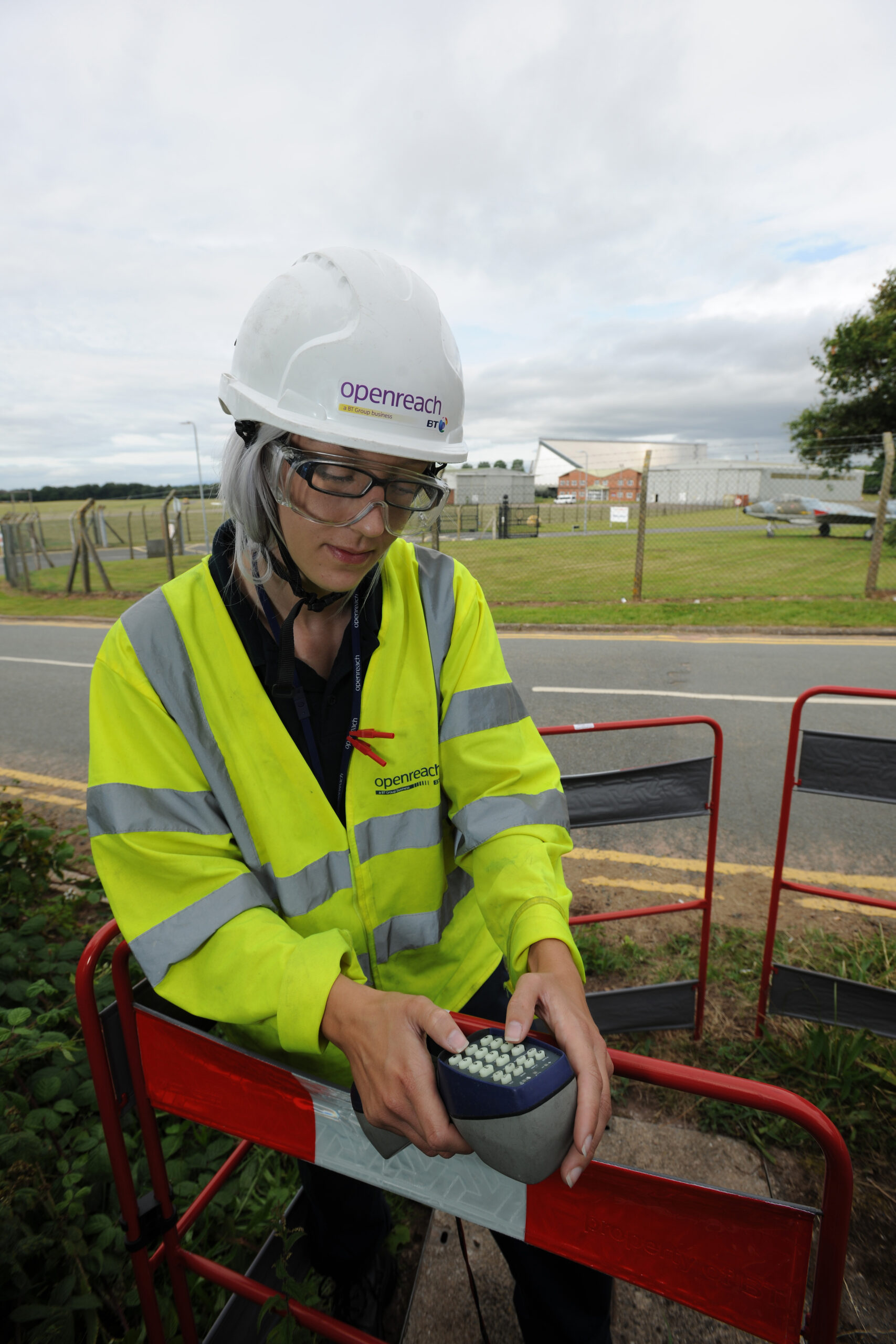 A woman wearing a hard hat, goggles and a hi vis vest in using a hand held monitoring device, whilst rounded by safety fencing. In the background are aircraft. Open Reach Logo is visible on her vest